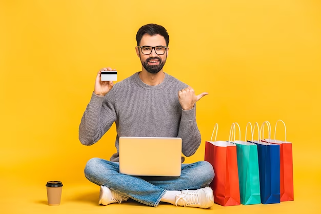 A photo of a man sitting in front of a computer, holding up a credit card with shopping bags next to him.